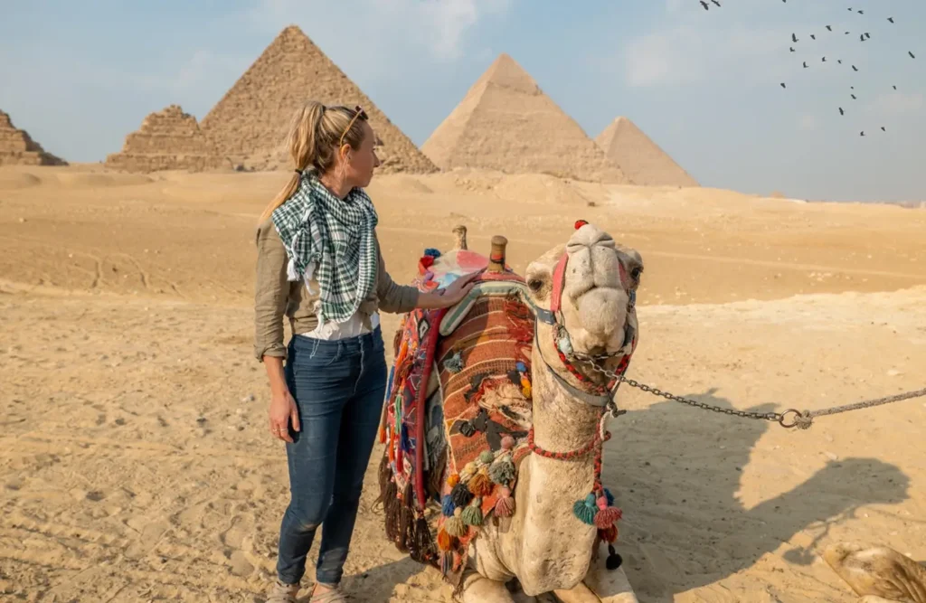 A visitor standing beside a camel in front of the pyramids